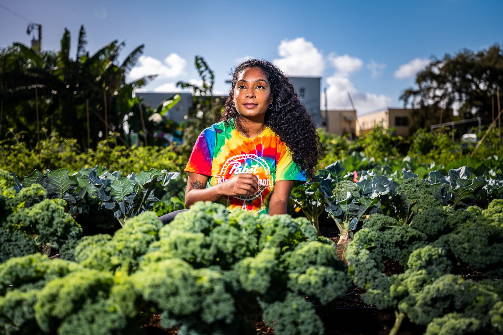 A woman in a tie-dye shirt stands among rows of kale in a sunlit Miami community garden